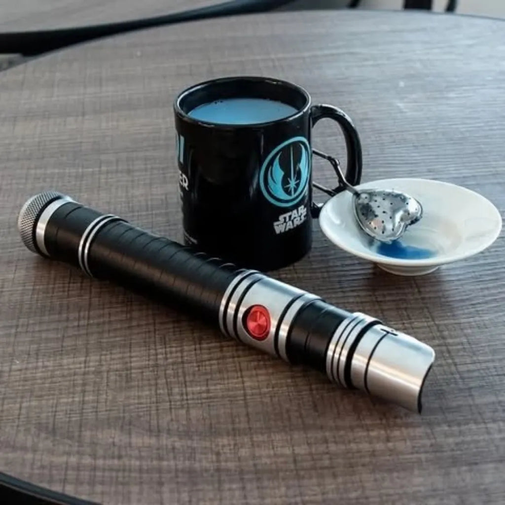 A black and silver lightsaber on a wooden table next to a mug of blue milk