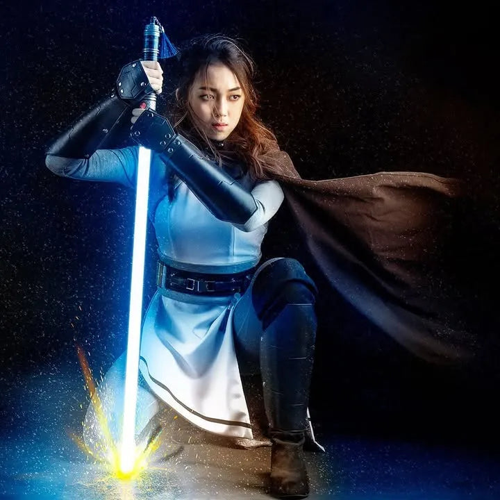 Female Jedi cosplayer kneeling in a defensive stance, stabbing a blue lightsaber into the ground amidst flying sparks