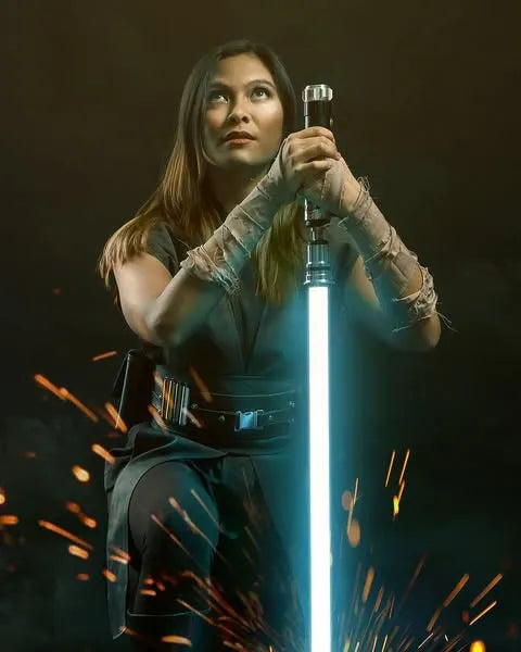 Female Rey-inspired cosplayer kneeling and holding a Luke Skywalker-inspired custom neopixel lightsaber with a light blue blade