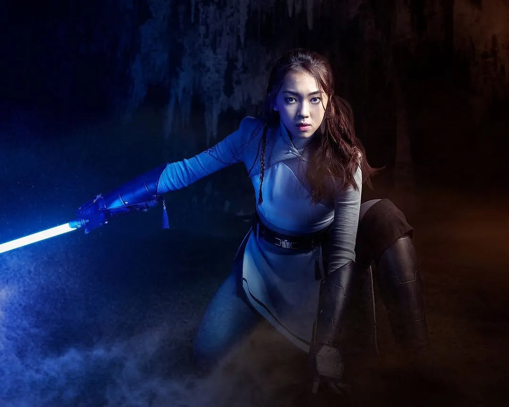 Female Jedi-inspired cosplayer kneeling in a defensive stance holding a Rahm Kota-inspired custom neopixel lightsaber with a blue blade
