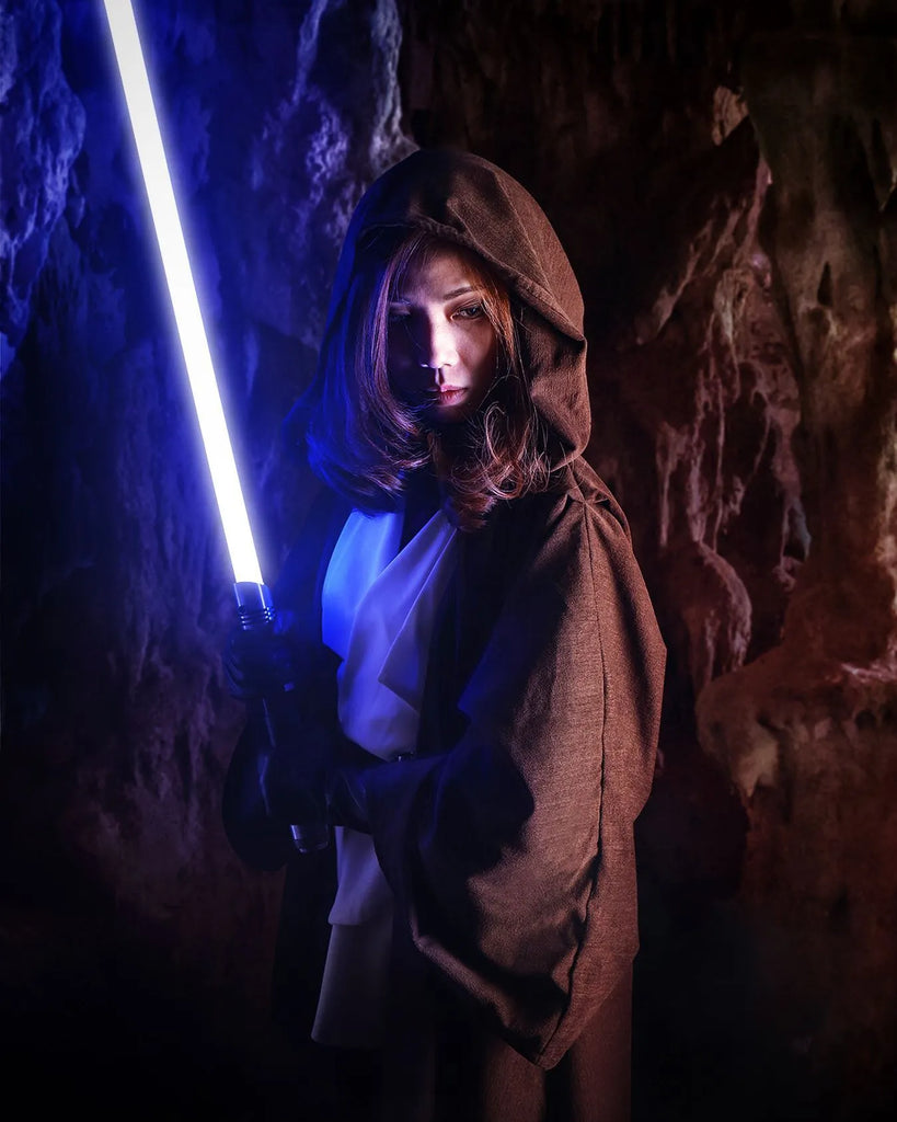 Female Jedi-inspired cosplayer standing in a dark cave holding a Darth Vader-inspired custom neopixel lightsaber, illuminated with bright blue blade