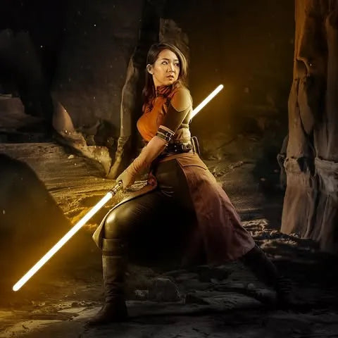 Female Jedi-inspired cosplayer kneeling in a defensive stance inside a cave wielding saberstaffed Arclyte with yellow blades